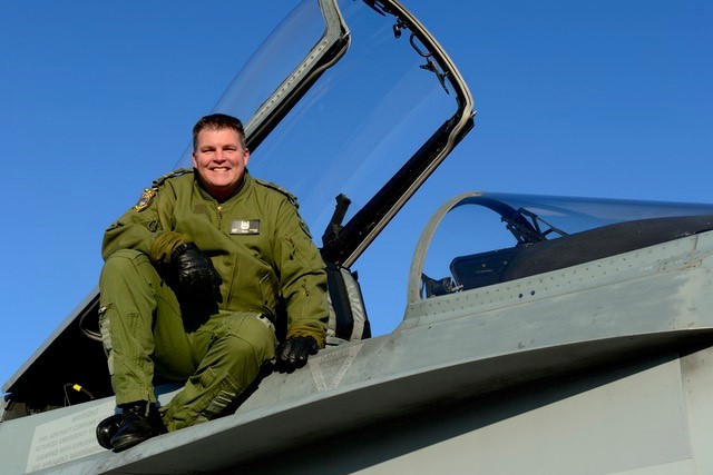 A military member in uniform is sitting on top of a fighter jet with an open cockpit. He is smiling at the camera.