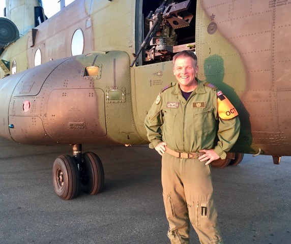 A military member in uniform is standing in front of a Chinook helicopter while smiling at the camera.