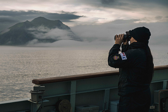 Person using binoculars on a ship deck, overlooking misty mountains across calm water.