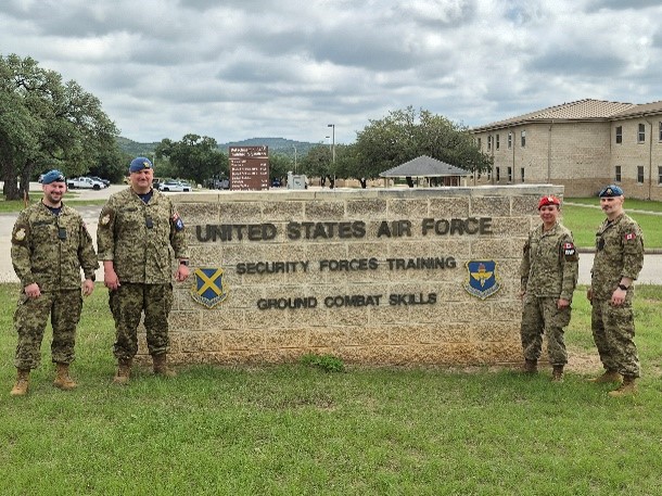 Four people in camouflage military uniforms stand on grass on either side of a large stone sign that reads “United States Air Force Security Forces Training Ground Combat Skills.” 
