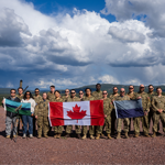 A large group of uniformed military personnel stands outdoors on rocky ground, posing in a line while holding three flags, including a Canadian flag in the center. Behind them are rolling hills and a distant wind farm, under a dramatic sky filled with large clouds and patches of blue.