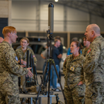 Several uniformed military personnel stand around a table indoors, examining and discussing electronic equipment mounted on a tripod. A tall antenna-like device is positioned at the center of the setup. Additional people and vehicles are visible in the blurred background of the large hangar-like space.