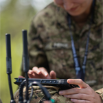 Close-up of a uniformed individual outdoors using a handheld device, with radio antennas visible in the foreground and a blurred natural background.