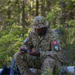 Uniformed individual sits on a fallen log in a forest, using a handheld device beside a bag and equipment, surrounded by trees and greenery.