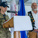 A Canadian Armed Forces member in camouflage uniform stands at a podium speaking into a microphone during an event. Behind the podium, another person wearing traditional regalia stands near several flags and ceremonial decorations inside a tented venue.