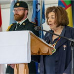 An individual wearing a commemorative medallion stands at a podium speaking into a microphone during a ceremony. Several flags are displayed in the background, and uniformed military personnel stand nearby on a stage inside a tented venue.