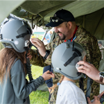 Two military members stand beside two children under a tent, helping them try on aviation-style helmets. One child is already wearing a helmet while the other is having a helmet adjusted.