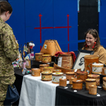 A person in a Canadian Armed Forces camouflage uniform stands at a display table showcasing handcrafted items, including decorated birch bark baskets, containers, and artwork. Two vendors sit behind the table in a gymnasium-like venue, with numerous handmade pieces arranged on black and white tablecloths.