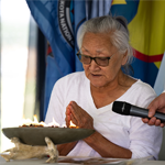 An individual sits at a table with hands clasped in front of a shallow ceremonial bowl containing a small flame. A microphone is held nearby by another participant, while several flags and part of a traditional regalia garment are visible in the background inside a tented venue. A small animal skull and other ceremonial items rest on the table beside the bowl.