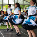 Three dancers perform on a stage beneath an event tent, wearing coordinated blue, white, and black ruffled costumes with matching footwear. The dancers are captured mid-step with skirts lifted as part of the routine. Audience members seated at tables watch from the background.
