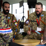 Several participants sit in a circle around a large drum during a cultural performance inside a tented venue. Two Canadian Armed Forces members in camouflage uniforms hold drumsticks and strike the drum in unison, while other participants seated around the drum also take part. Flags, event signage, and additional attendees are visible in the background.