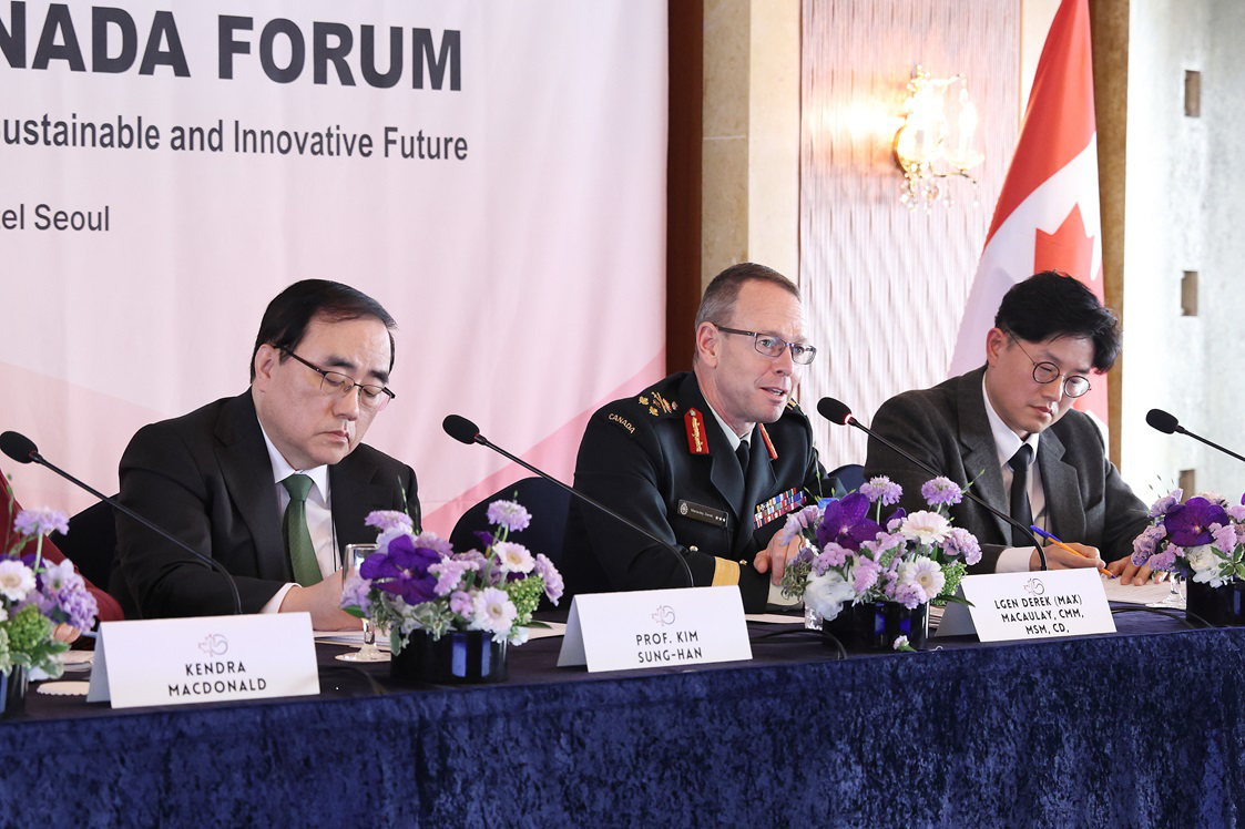 Lieutenant-General Derek Macaulay speaks during a panel discussion at the 22nd Korea–Canada Forum in Seoul, South Korea. Seated alongside fellow panelists at a conference table with microphones and floral arrangements, he addresses attendees with a Canadian flag displayed behind the panel.
