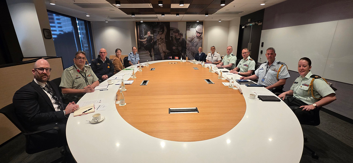 Lieutenant-General Derek Macaulay meets with senior leadership from the Ministry of Defence and Defence Force around a large conference table during a briefing session. Participants discuss regional and global security issues in a modern meeting room with briefing materials, notebooks and refreshments on the table.