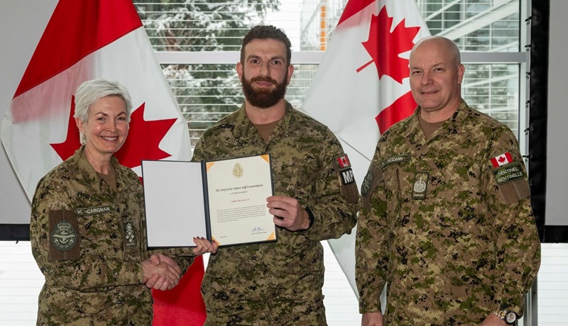 Canadian Armed Forces member stands between two uniformed members, holding an award certificate in front of two Canadian flags.