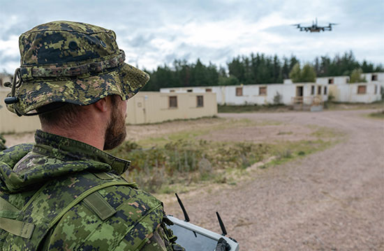 Military personnel operating a controller while monitoring a drone over a training area.
