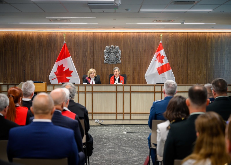 Audience members sit facing a judicial bench in a formal courtroom setting. The Chief Military Judge and the Chief Justice of the Court Martial Appeal Court of Canada are wearing red and black judicial robes and are seated beneath the Canadian coat of arms, with Canadian flags displayed on either side of the bench.