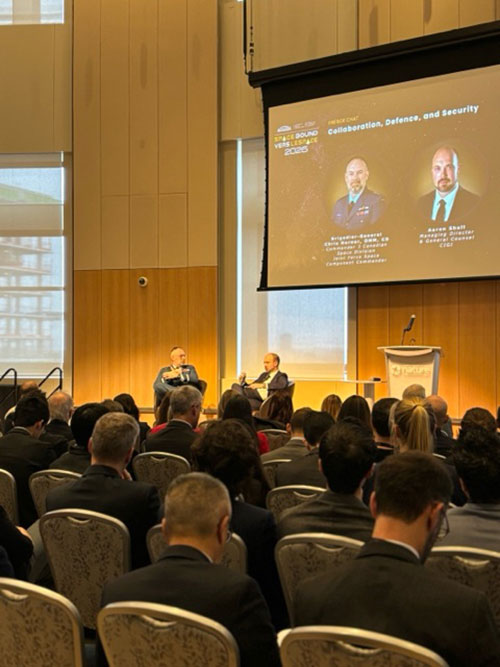 A seated audience watches two presenters seated on a stage and a screen in the background features the names and titles of the presenters.