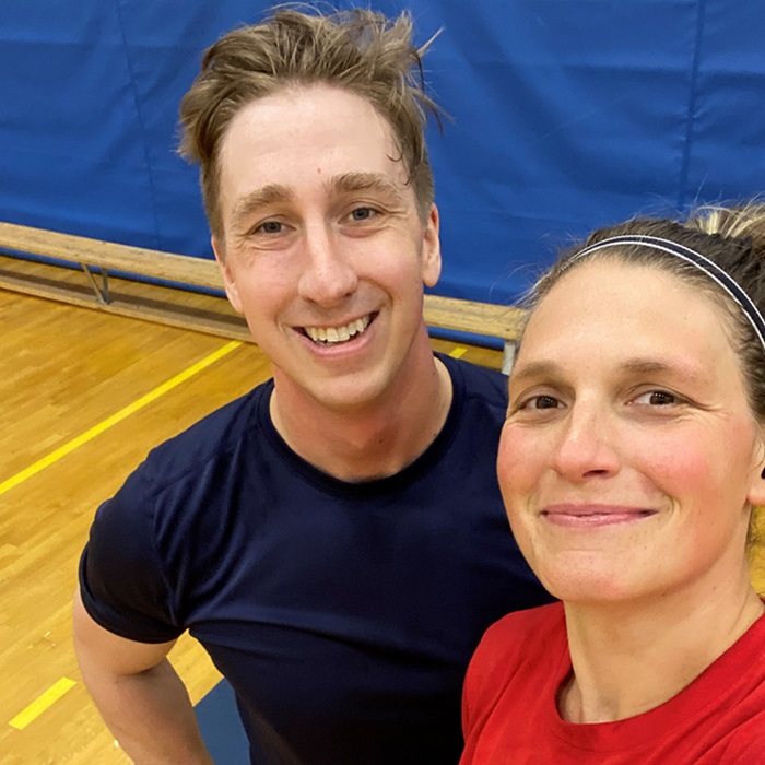 Un égoportrait en gros plan de deux personnes dans un gymnase. À l'arrière-plan, un plancher de bois franc avec des lignes jaunes et un mur capitonné bleu.