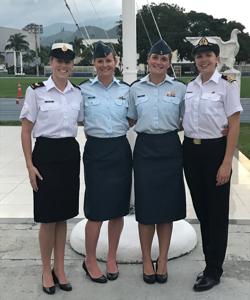 Quatre femmes en uniformes de style militaire se tiennent à l'extérieur, devant un mât de drapeau. Deux portent des chemises bleu pâle et des jupes foncées, et deux portent des chemises blanches et des jupes ou pantalons foncés. Elles portent toutes une casquette ou un béret et des chaussures noires.