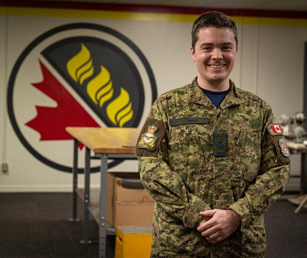 Une personne vêtue d’un uniforme militaire est debout à l’intérieur. Derrière elle se trouve un grand emblème mural représentant une feuille d’érable rouge stylisée, trois flammes jaunes et une bordure noire circulaire. La pièce, dont les murs sont peints d’une couleur neutre, contient des tables, des boîtes et du matériel.