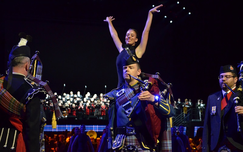 Master Corporal Tim Reid, Pipe Instructor for the Royal Canadian Air Force Pipes and Drums, stands with his bagpipes in hand during the opening performance of the Virginia International Tattoo in Norfolk, Virginia at the Norfolk Scope Arena on 23 April 2025. A highland dancer on a podium behind him strikes a pose. Dancers and military musicians on the floor, in addition to a choir on risers can be seen in the background.