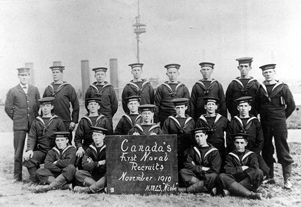 Nineteen men pose with a sign which reads Canada’s First Naval Recruits November 1910, HMCS Niobe.
