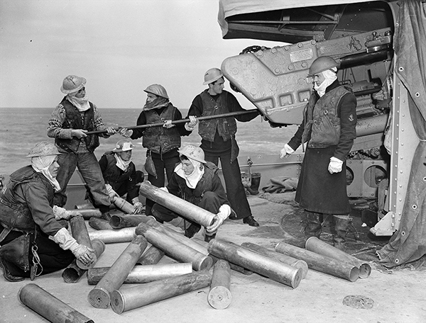 Three Naval officers use a rod to clean out a large gun. Four other officers inspect the gun’s shells which have been laid out on the sand.