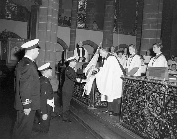 A Naval officers hands a flag over to a priest. Members of the church and Navy stand as witnesses.