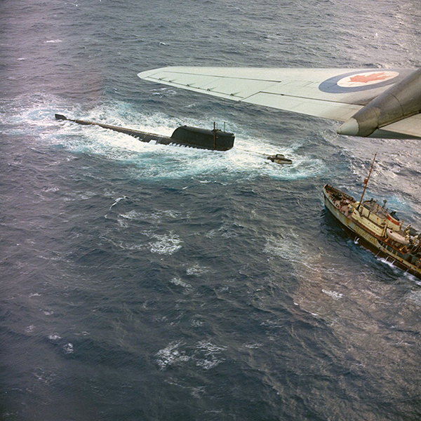 A submarine surfaces on the water. Another ship approaches it while a Canadian aircraft flies overhead.