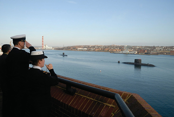 Three Naval officers watch as two submarines pass them in the water. One officer salutes, as another plays the pipe.