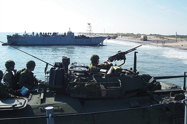 Soldiers in an armoured vehicle watch as a naval ship approaches the shore.