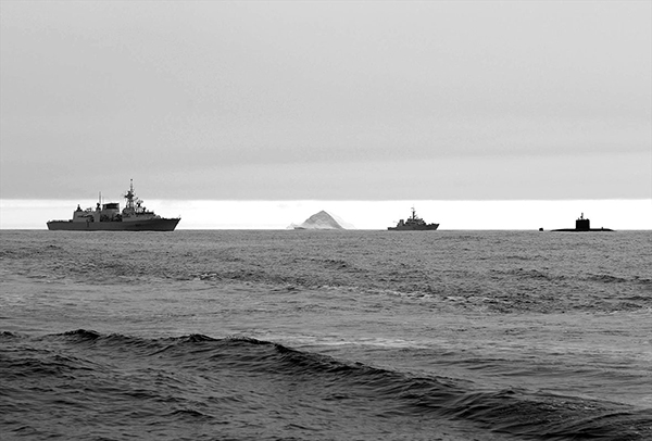 Two ships and a submarine pass an iceberg on the water.