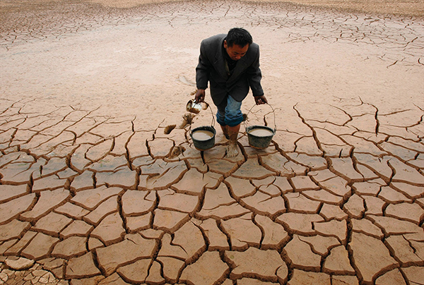 A man carries a bucket of water in each hand across dry, cracked land.