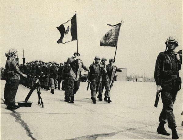 Soldiers in formation, parading, with 2 flags.