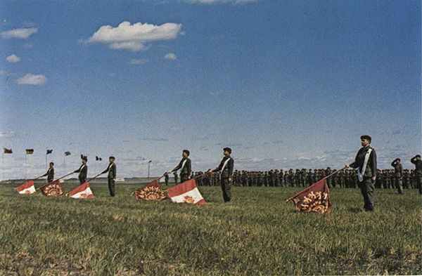 Soldiers bearing lowered flags, at attention in a field.