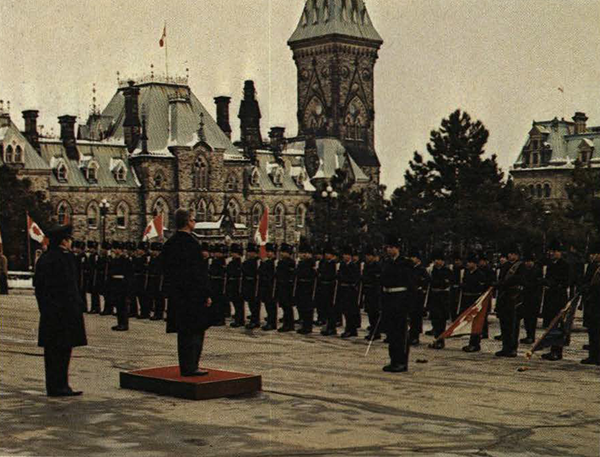 Man on a podium in front of soldiers at attention, Parliament in background
