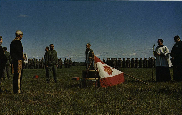 Soldiers, at ease, in a field, with the flag leaning on drums. A priest in attendance.