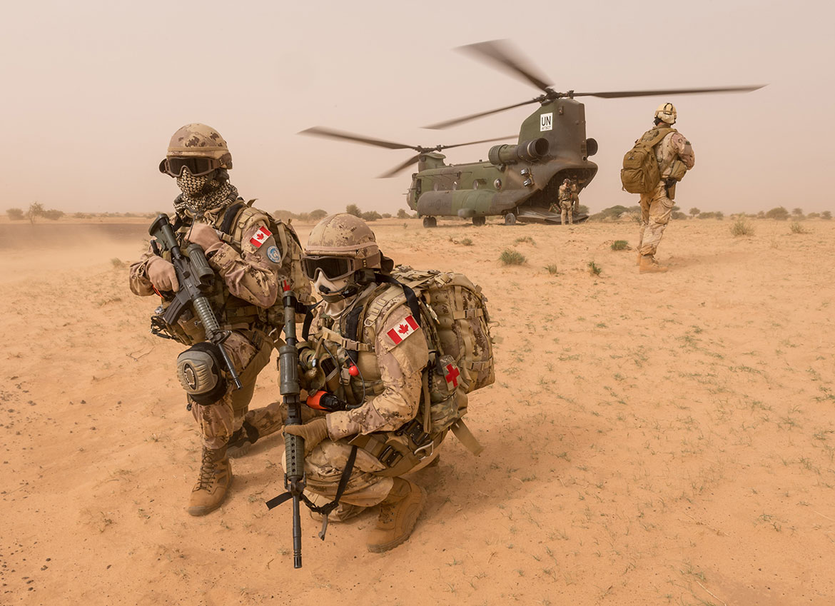 Two Canadian soldiers look out into the distance as other soldiers exit an aircraft.