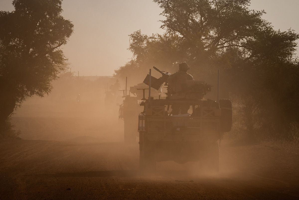 Military vehicles drive in a line down a dusty African road between trees. Military members are seated on mounted weaponry on the back of the vehicles. The image is cloudy due to all the dust kicked up into the air by the vehicles.