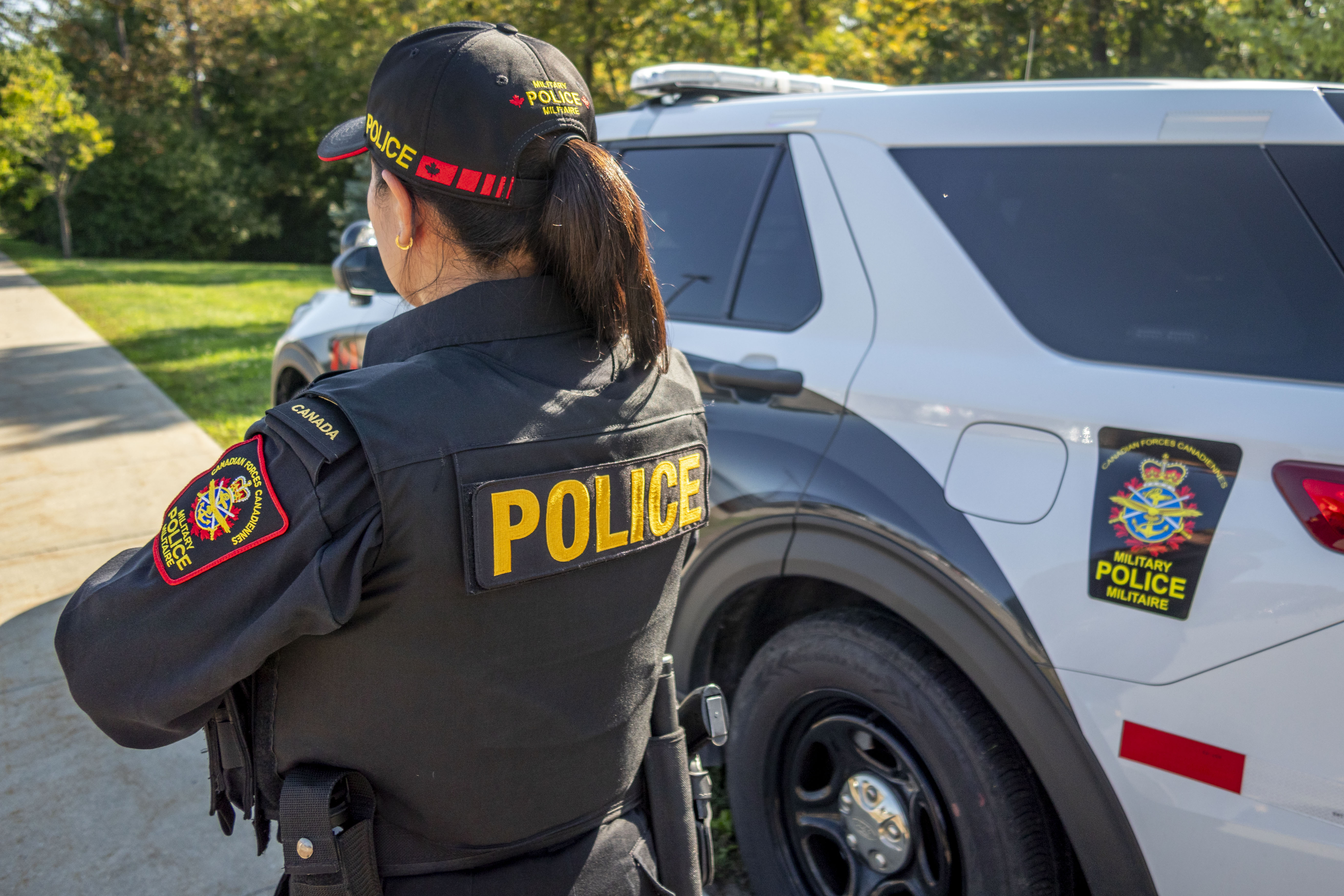 A member of the Military Police is shown wearing the
military police ball cap from behind. The female
member is also wearing a police vest and black uniform
with POLICE written in capital letters in yellow. A
white and black military police vehicle is in the
background with trees.