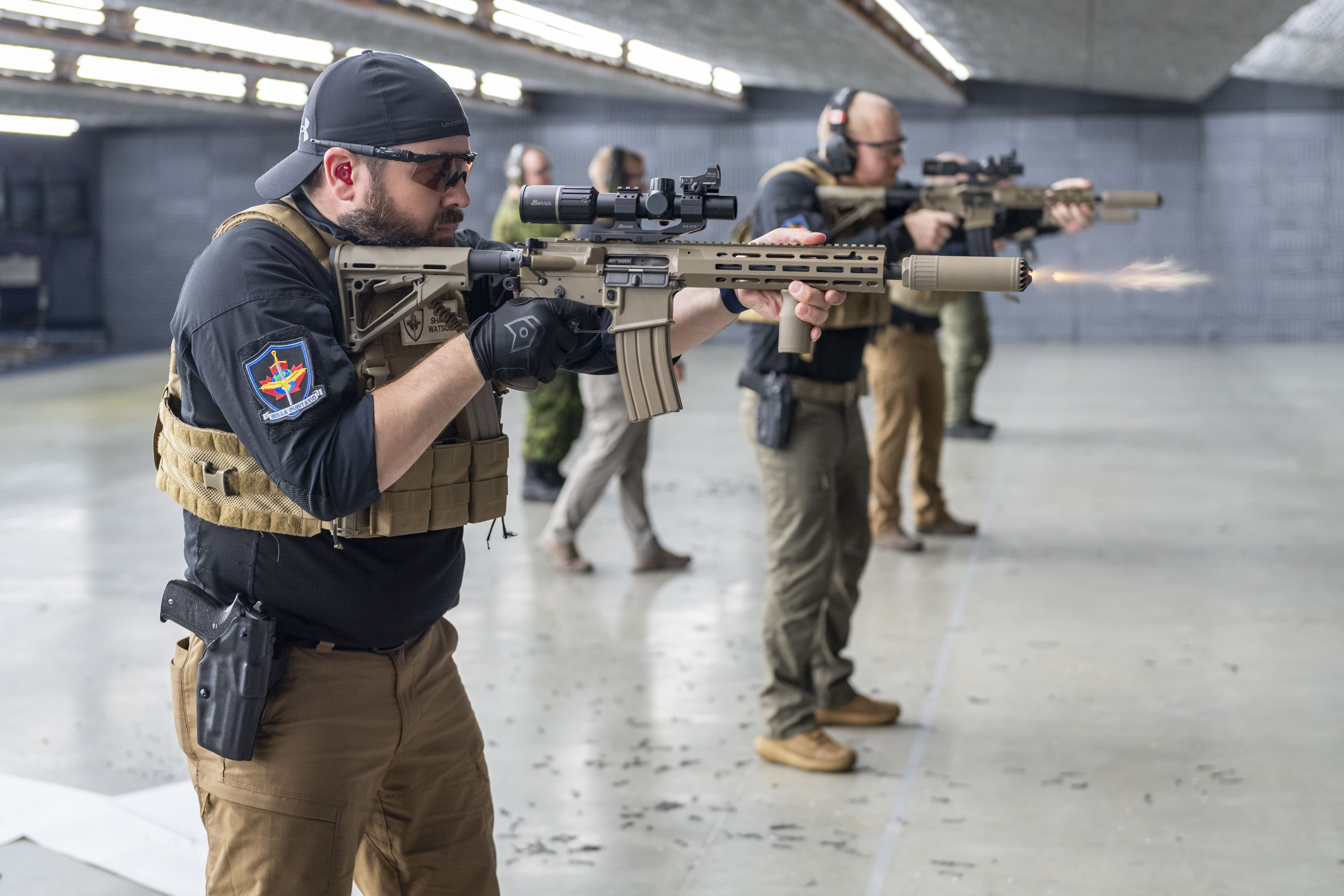 Aircraft Security Officers stand in a line on an indoor firing range with brown rifles aimed in
front of them with pistols on their belts. A burst of flame is coming out of the nearest rifle.