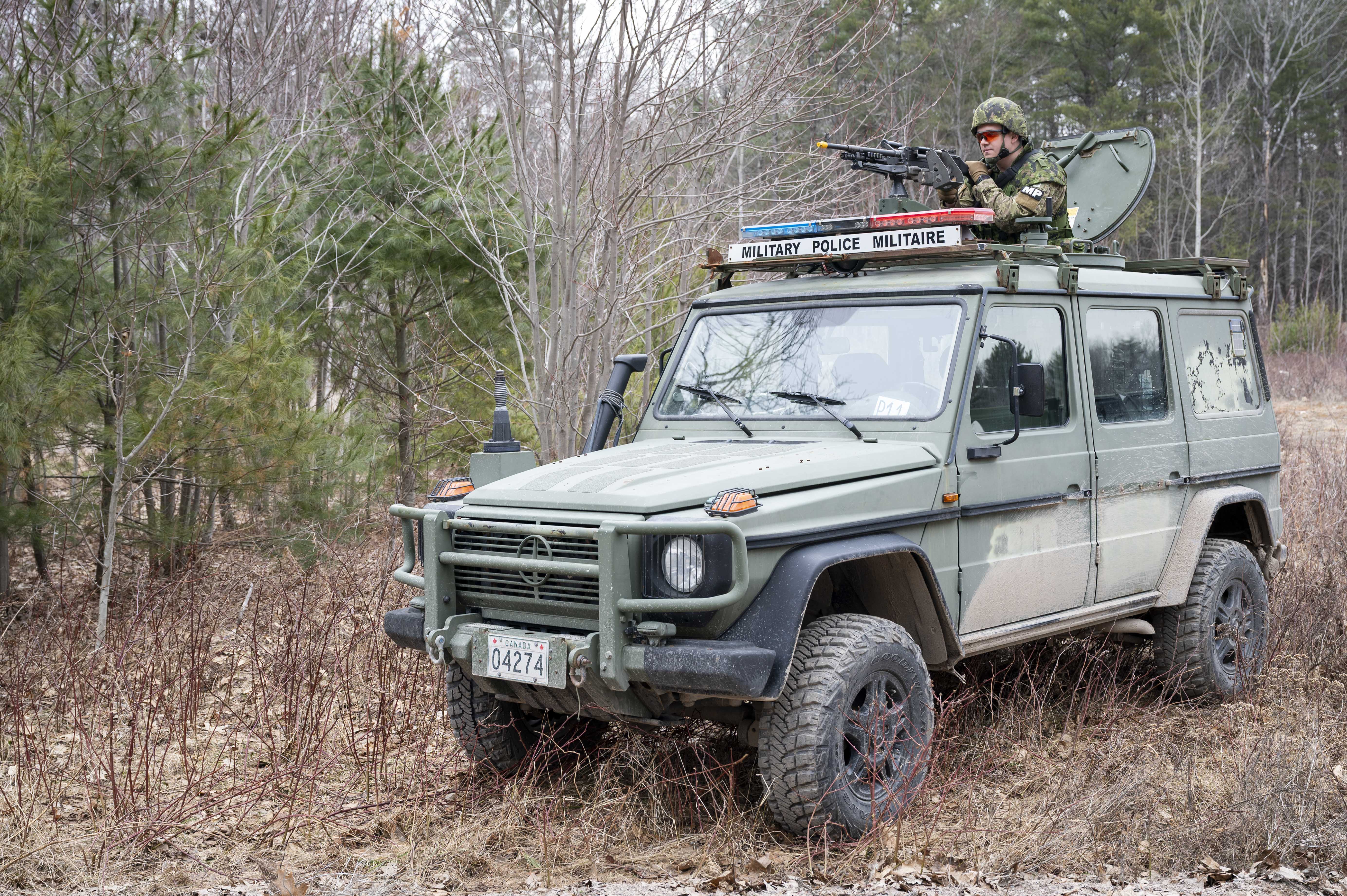 A green Military Police vehicle with the words “Military Police Militaire” is parked in the forest
with a Military Police member standing out of the vehicle roof harch aiming his firearm.