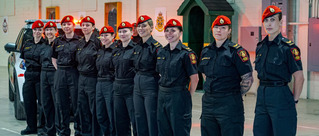 Female Military Police members stand in a line wearing their black uniform and the distincitve
red beret of the Military Police. Behind them, a Military Police car has its lights flashing.