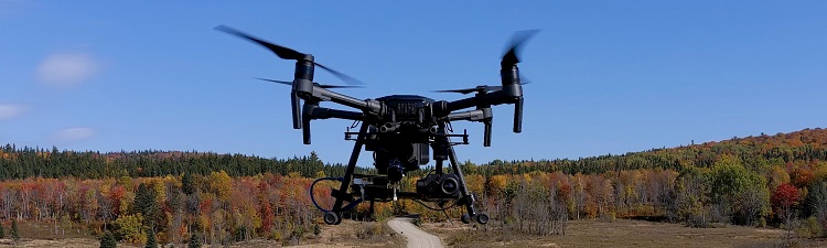 A black quad-copter drone flies against a blue sky. Trees are in autumn colours in the background.