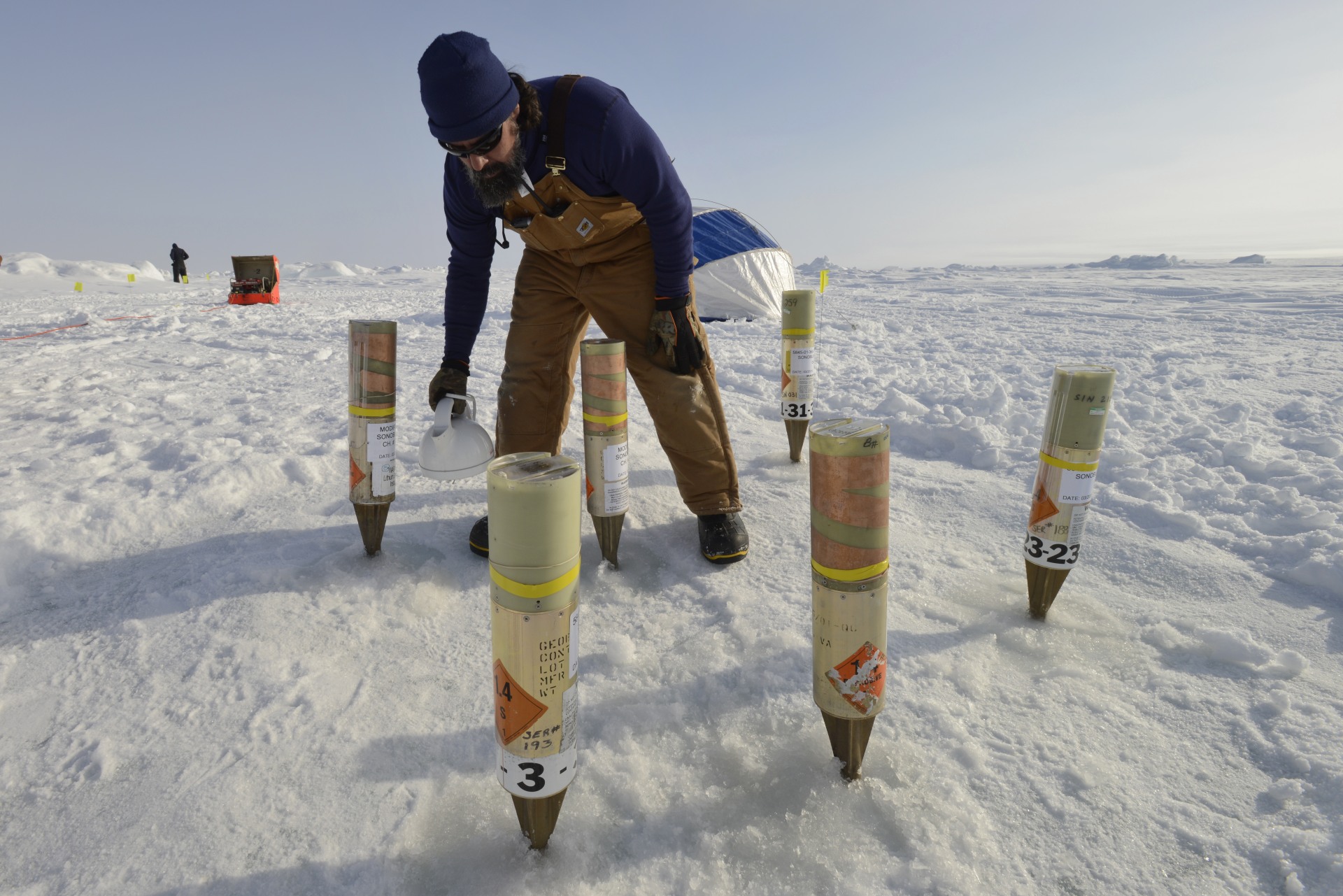 Watering a geobuoy garden. The buoys were inserted with the entire icepick embedded in the holes, then a slurry of ice and water was poured around the holes and allowed to freeze, coupling the geobuoys to the ice in a controlled manner.
Photo by Janice Lang, DRDC 
