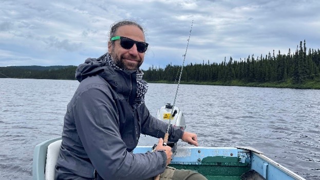 Un homme souriant portant un foulard, un manteau et des lunettes de soleil, assise dans une chaloupe sur un lac avec une canne à pêche.