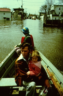 Photo - Immeasureable loss. A man and a child riding in a boat surrounded by buildings submerged in water.