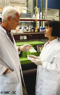 Un homme et une femme scientifiques se tiennent côte à côte pour discuter d’un échantillon de laboratoire devant leur équipement de laboratoire.
