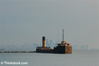Un vieux navire est amarré près d’un mur de roches. Au loin, la silhouette de la ville de Toronto est masquée par une couverture gris sombre.