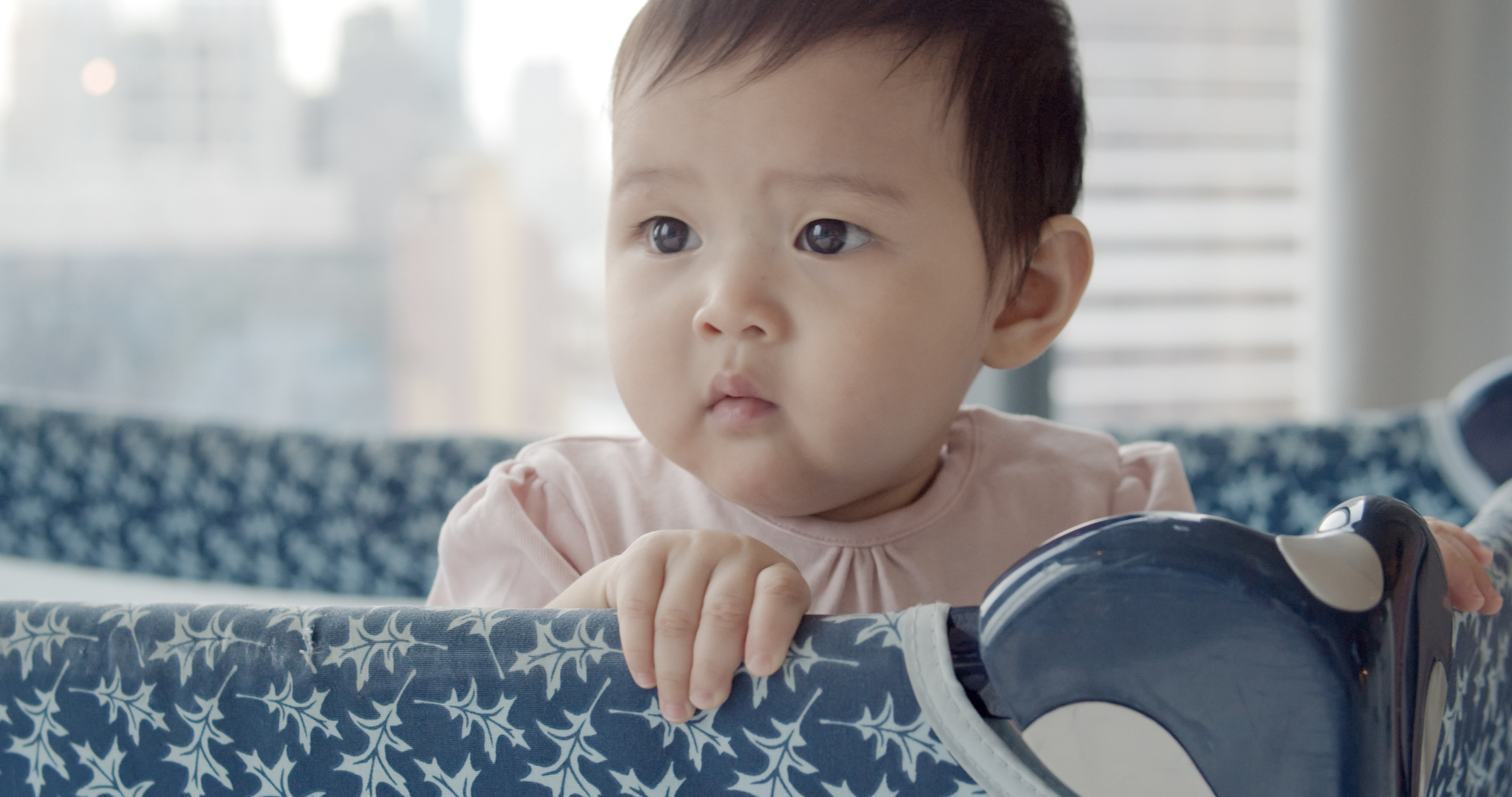 A baby stands and looks over the side of a playpen holding on with both hands.